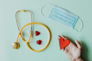 Top view of crop anonymous person hand with red paper heart on table with stethoscope and medical mask for coronavirus prevention